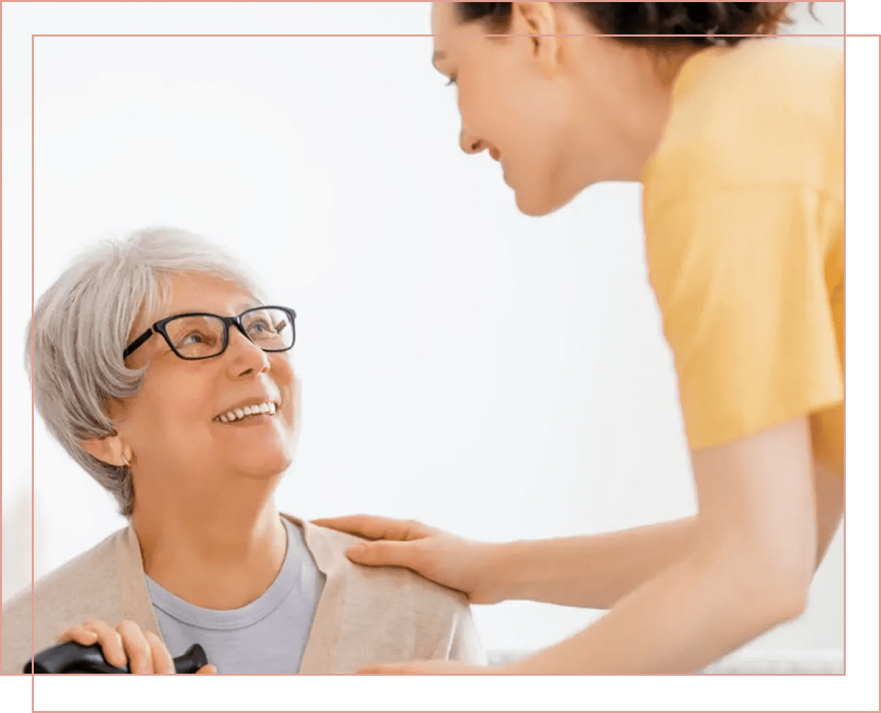 A caregiver warmly interacts with an elderly woman wearing glasses.